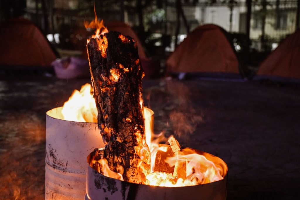 Glowing campfire in a barrel among tents, creating a warm nighttime ambiance.