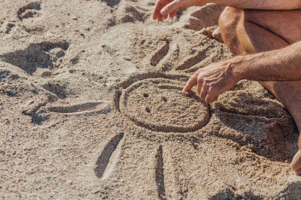 Hands drawing a cheerful sun in the sand, capturing the essence of a sunny beach day.